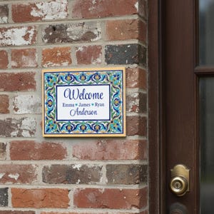 K&ouml;nnte beinhalten: Rechteckiges Willkommensschild mit dekorativem Rand in Blau-, Gr&uuml;n- und Gelbt&ouml;nen. Auf dem Schild steht "Welcome" &uuml;ber den Namen "Emma James Ryan Anderson" in einer Schreibschrift. Das Schild ist an einer Backsteinmauer angebracht.