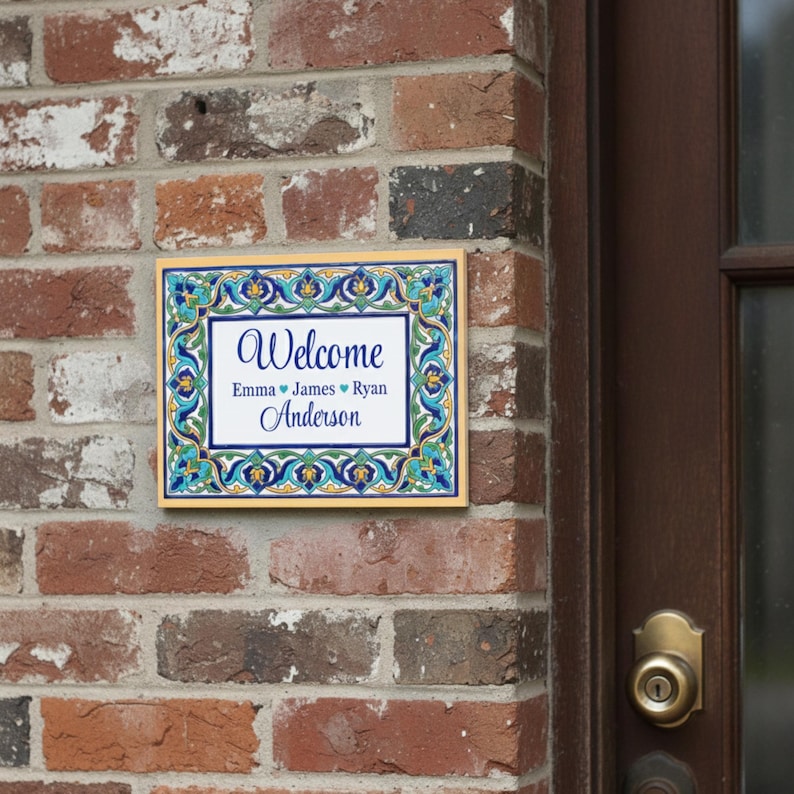 K&ouml;nnte beinhalten: Rechteckiges Willkommensschild mit dekorativem Rand in Blau-, Gr&uuml;n- und Gelbt&ouml;nen. Auf dem Schild steht "Welcome" &uuml;ber den Namen "Emma James Ryan Anderson" in einer Schreibschrift. Das Schild ist an einer Backsteinmauer angebracht.
