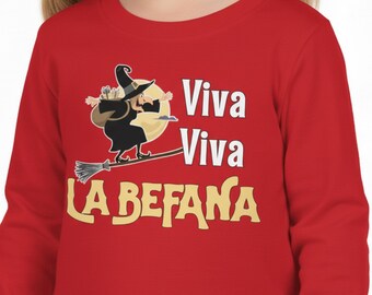 Puede incluir: Una camiseta roja de manga larga con un dibujo animado de una bruja en una escoba frente a una luna llena. El texto "Viva Viva LA BEFANA" está en blanco y dorado. La bruja lleva un sombrero y una capa negros. Un diseño festivo.
