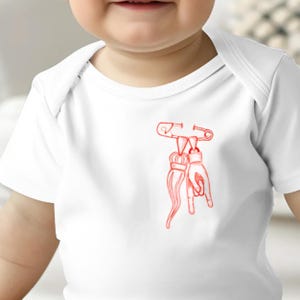 May include: A white baby onesie with a red illustration of a safety pin holding two objects, one resembling a hand giving the rock and roll sign and the other a long, thin object with a crown at the top.
