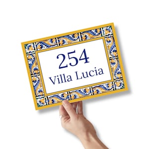 May include: A rectangular sign with the number "254" and the words "Villa Lucia" in navy blue. The sign has a white center with a decorative border featuring blue, yellow, and gold floral patterns. The sign is held by a hand.