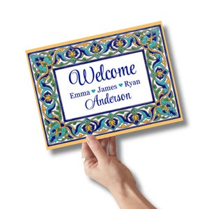 K&ouml;nnte beinhalten: Rechteckiges Willkommensschild mit dekorativem Rand in Blau-, Gr&uuml;n- und Goldt&ouml;nen. Das Schild tr&auml;gt das Wort "Welcome" in eleganter Schrift sowie Namen und einen Nachnamen. Das Schild wird von einer Hand gehalten.