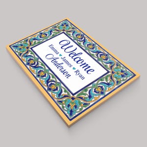 K&ouml;nnte beinhalten: Rechteckiges Willkommensschild mit dekorativem Rand in Blau-, T&uuml;rkis- und Gelbt&ouml;nen. Das Schild zeigt das Wort "Welcome" in eleganter Schrift sowie Namen. Das Schild hat eine wei&szlig;e Mitte und einen gelben Rand.
