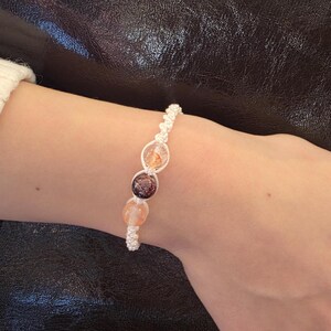 May include: A white string bracelet with three round beads. The beads are clear with a light pink, brown, and orange colour.