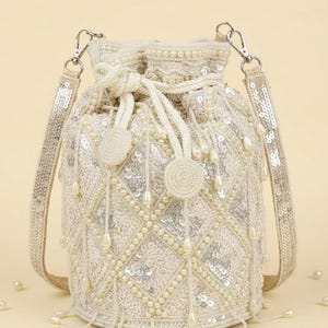 Puede incluir: Un bolso tipo saco de color crema con un patrón de diamantes creado con cuentas de perlas blancas y lentejuelas plateadas. El bolso incluye un cierre de cordón con dos borlas de cuentas, una correa de lentejuelas y adornos de perlas colgantes.