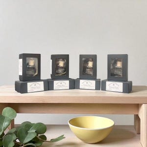 May include: Four boxed car air fresheners displayed on a wooden surface. Each dark grey box has a clear window revealing a glass bottle with a wooden top. The boxes are labelled with scent names: Tobacco Grove, Brown Blue, and Cotton Mist.