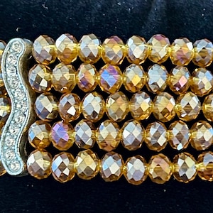 May include: A multi-strand bracelet featuring five rows of faceted amber-colored glass beads. The rows are held together by two vertical silver-toned metal bars, each inlaid with a single column of small, clear, sparkling rhinestones.
