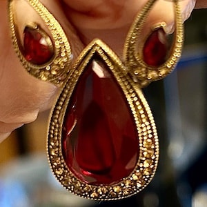 May include: A gold-toned pendant necklace featuring a large, central, pear-shaped deep red gemstone surrounded by a textured, beaded metal border. Two smaller, matching pear-shaped red gemstones are set in ornate, curved gold-toned metal frames.