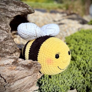 May include: A crocheted bee toy with black and yellow stripes, white wings, and a smiling face. The bee is sitting on a piece of driftwood and green moss.
