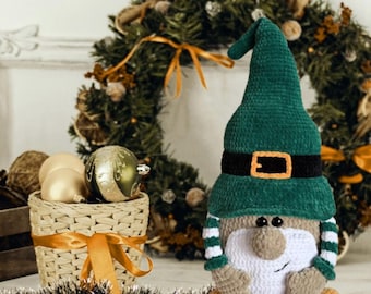 May include: A handmade crochet gnome with a green hat, black belt, and orange feet sits in front of a Christmas wreath. A basket with gold ornaments and a garland are also in the image. The gnome is a festive holiday decoration.