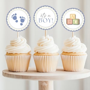 May include: Three cupcakes with blue and white frosting topped with round paper cupcake toppers. The toppers feature a baby footprint, the words "it's a BOY!" and a stack of alphabet blocks.