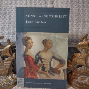 May include: A copy of "Sense and Sensibility" by Jane Austen. The book has a teal cover with the title and author's name in white. The cover features a painting of two women in period clothing. The Barnes & Noble Classics logo is visible.