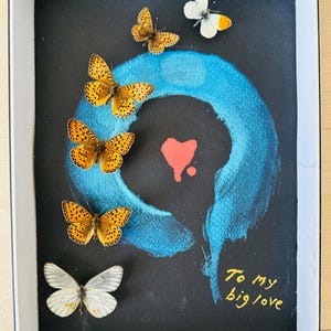 Pode incluir: Um fundo preto com um círculo azul pintado no centro. Há várias borboletas presas ao fundo, incluindo uma borboleta branca com marcas laranja. O texto "To my big love" está escrito em tinta dourada no canto inferior direito.