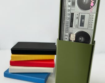 Puede incluir: Un porta tarjetas verde con una tarjeta de crédito Discover en su interior. La tarjeta presenta un diseño de radiocasete retro con la palabra "Discover" impresa en rosa. El porta tarjetas está colocado encima de una pila de cinco cuadernos de colores.