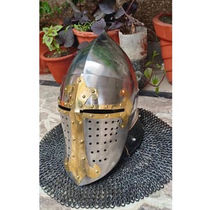 May include: A polished steel and brass medieval helmet with a cross design on the faceplate. The helmet features a riveted construction and is set on a chainmail coif. The brass accents add a decorative touch to the historical armour.