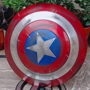 May include: A Captain America shield, featuring a red, white, and blue color scheme. The shield has a circular design with a silver star in the center. The shield is resting on a silver Avengers logo.