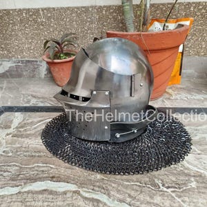 May include: A polished steel helmet with a visor and a chainmail neck guard. The helmet is sitting on a patterned surface. The text "TheHelmetCollection" is visible on the helmet.
