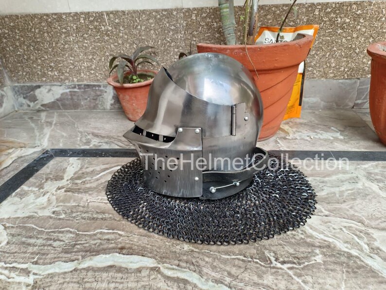 May include: A polished steel helmet with a visor and a chainmail neck guard. The helmet is sitting on a patterned surface. The text "TheHelmetCollection" is visible on the helmet.