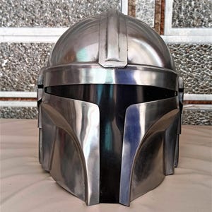 May include: A metallic silver helmet with a black visor. The helmet has a rounded top with a central ridge and angular features around the face and sides. The surface reflects light, creating highlights and shadows.