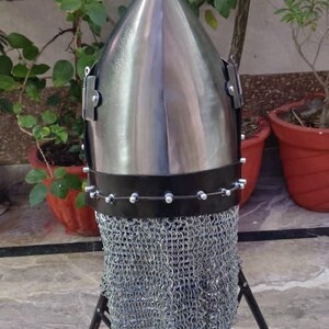 Peut inclure: Casque en m&eacute;tal poli avec un sommet conique, dot&eacute; d'une protection de cou en cotte de mailles. Le casque est mont&eacute; sur un support en m&eacute;tal noir. Le fond comprend des plantes vertes et des pots en terre cuite.