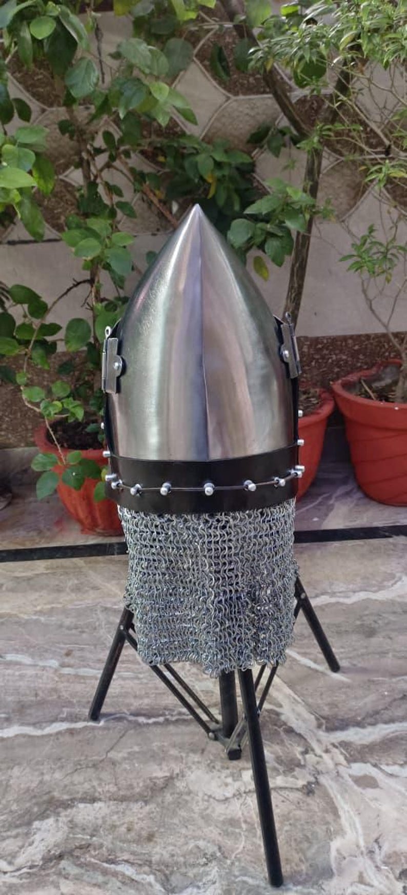 Peut inclure: Casque en m&eacute;tal poli avec un sommet conique, dot&eacute; d'une protection de cou en cotte de mailles. Le casque est mont&eacute; sur un support en m&eacute;tal noir. Le fond comprend des plantes vertes et des pots en terre cuite.