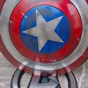 May include: A Captain America shield prop, featuring a red, white, and blue color scheme with a central white star on a blue background. The shield has a metallic silver border and visible battle damage. The Avengers logo is visible below.