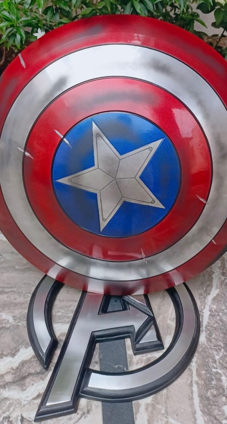 May include: A Captain America shield prop, featuring a red, white, and blue color scheme with a central white star on a blue background. The shield has a metallic silver border and visible battle damage. The Avengers logo is visible below.