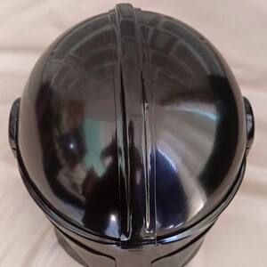 May include: A black, glossy helmet with a rounded top and a prominent central ridge. The helmet has a smooth, reflective surface and a visor-like shape at the front. The design includes ear-like protrusions on either side.