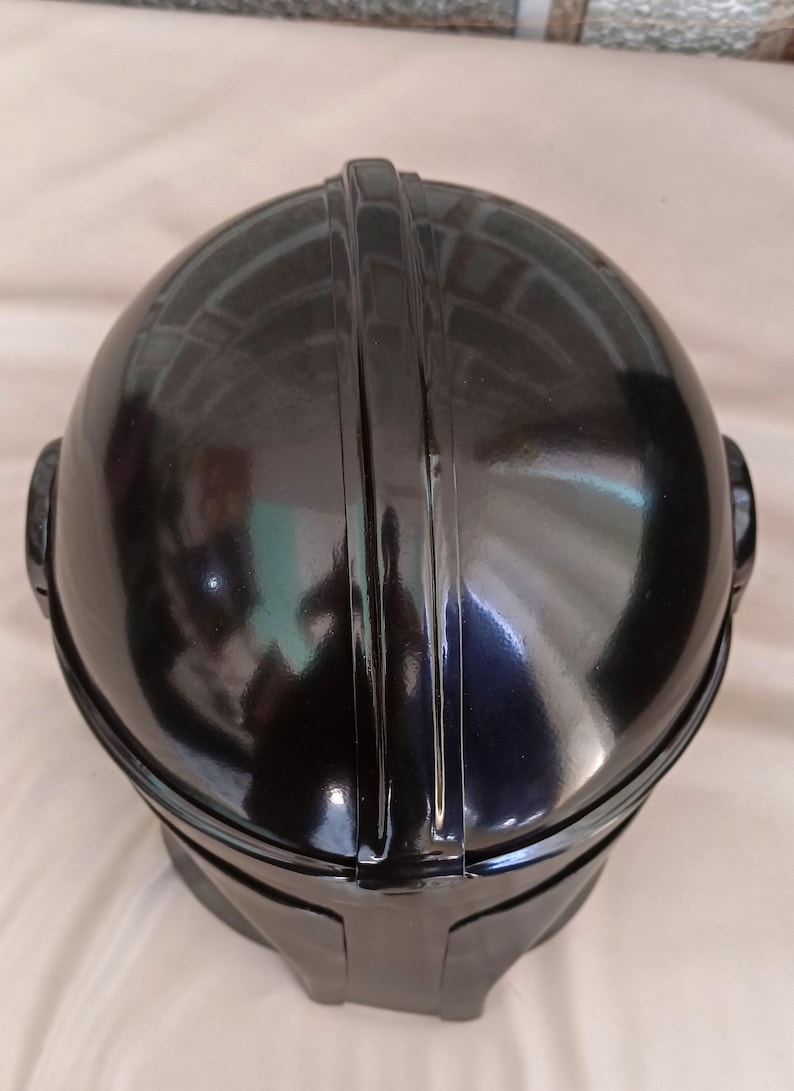May include: A black, glossy helmet with a rounded top and a prominent central ridge. The helmet has a smooth, reflective surface and a visor-like shape at the front. The design includes ear-like protrusions on either side.