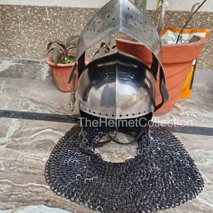 May include: A medieval-style helmet with a polished metal finish, featuring a visor with small perforations. The helmet includes a chainmail neck guard. The text "TheHelmetCollection" is visible on the helmet.