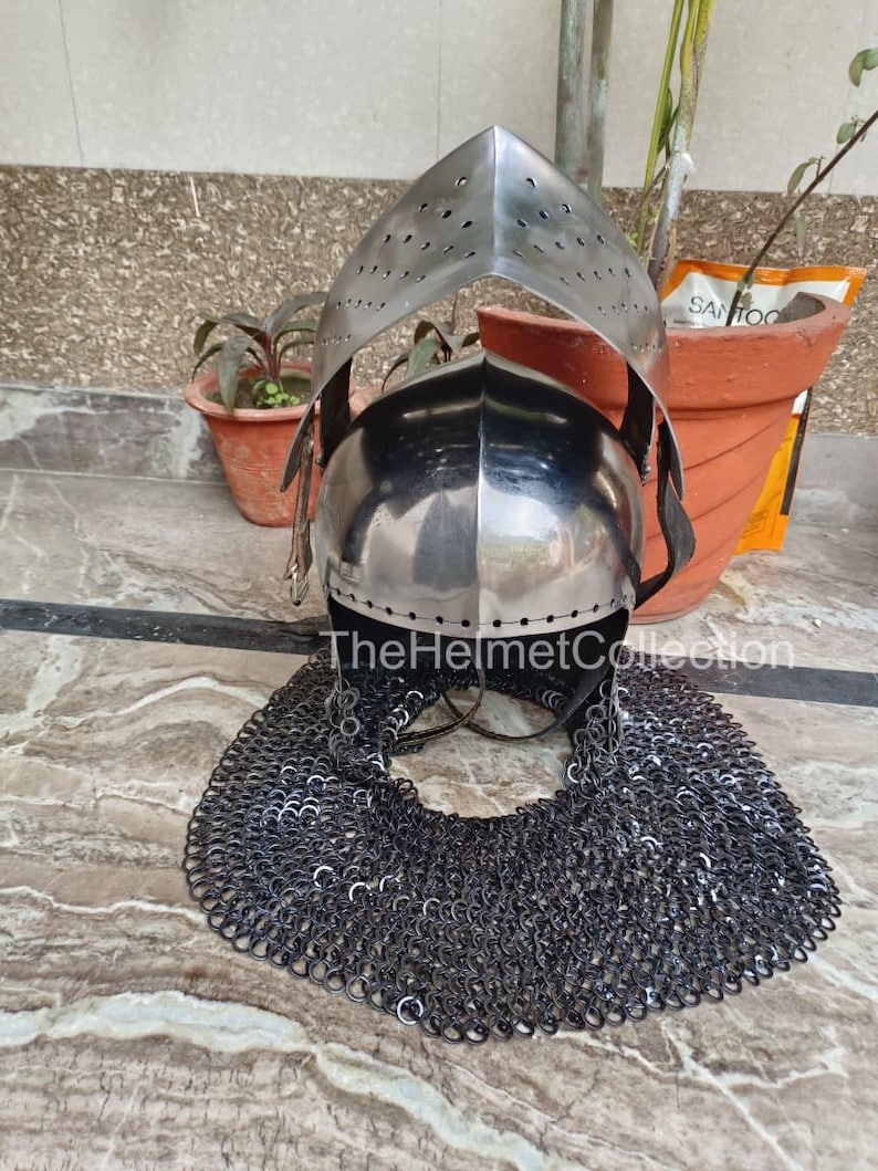 May include: A medieval-style helmet with a polished metal finish, featuring a visor with small perforations. The helmet includes a chainmail neck guard. The text "TheHelmetCollection" is visible on the helmet.