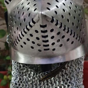 Peut inclure: Un casque m&eacute;di&eacute;val argent&eacute; avec une protection faciale perfor&eacute;e et une protection de cou en cotte de mailles. Le casque a un sommet pointu et une s&eacute;rie de petits trous espac&eacute;s. La cotte de mailles est faite de petits anneaux m&eacute;talliques.