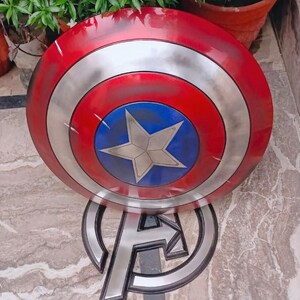 May include: A Captain America shield prop with a red, silver, and blue color scheme. The shield features a white star in the center of a blue circle, surrounded by concentric silver and red rings. The shield is resting on an Avengers logo.