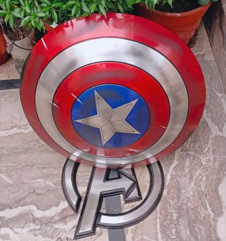 May include: A Captain America shield prop with a red, silver, and blue color scheme. The shield features a white star in the center of a blue circle, surrounded by concentric silver and red rings. The shield is resting on an Avengers logo.