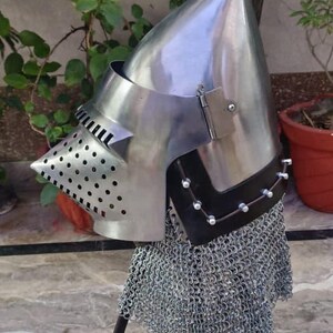 Peut inclure: Casque m&eacute;di&eacute;val en acier poli avec un sommet pointu, une visi&egrave;re perfor&eacute;e et une protection de cou en cotte de mailles. Le casque est expos&eacute; sur un support noir, mettant en valeur son design historique.