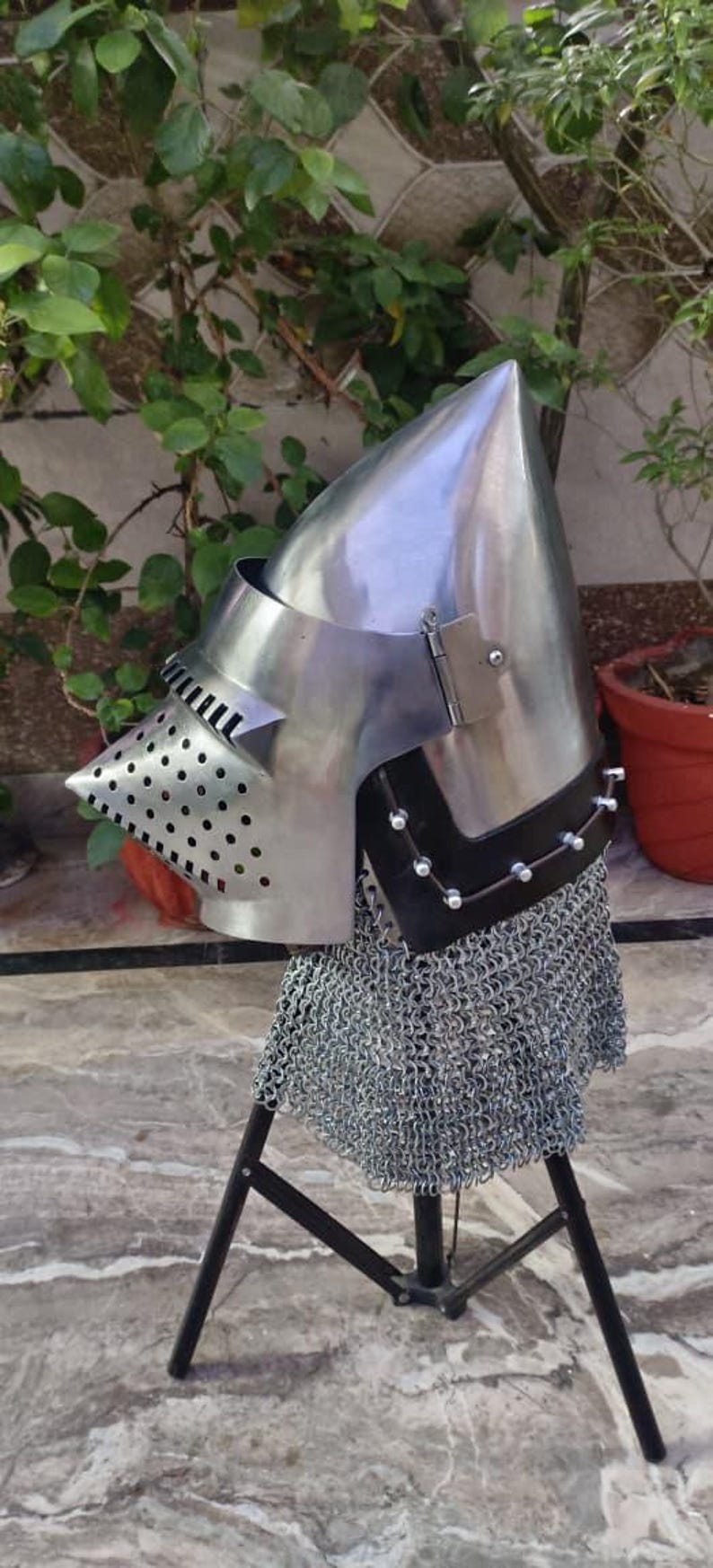 Peut inclure: Casque m&eacute;di&eacute;val en acier poli avec un sommet pointu, une visi&egrave;re perfor&eacute;e et une protection de cou en cotte de mailles. Le casque est expos&eacute; sur un support noir, mettant en valeur son design historique.
