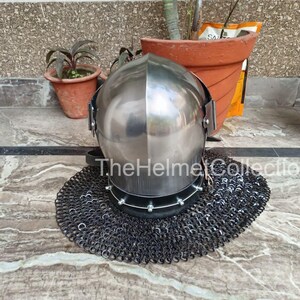 May include: A polished steel helmet with a rounded top, featuring a visor and riveted details. It rests on a circular chainmail coif. The text "The Helmet Collection" is visible on the helmet.