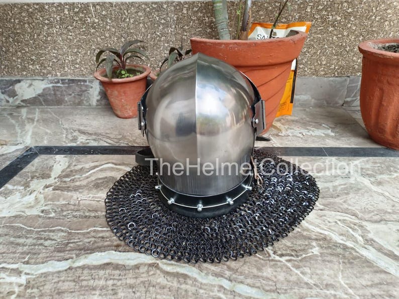 May include: A polished steel helmet with a rounded top, featuring a visor and riveted details. It rests on a circular chainmail coif. The text "The Helmet Collection" is visible on the helmet.