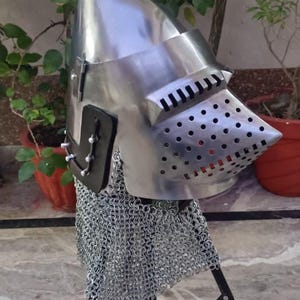 Peut inclure: Casque m&eacute;di&eacute;val en m&eacute;tal poli, avec une pointe et une visi&egrave;re perfor&eacute;e. Il est fix&eacute; &agrave; une protection de cou en cotte de mailles et est pr&eacute;sent&eacute; sur un tr&eacute;pied noir. Le fond comprend de la verdure et une surface carrel&eacute;e.