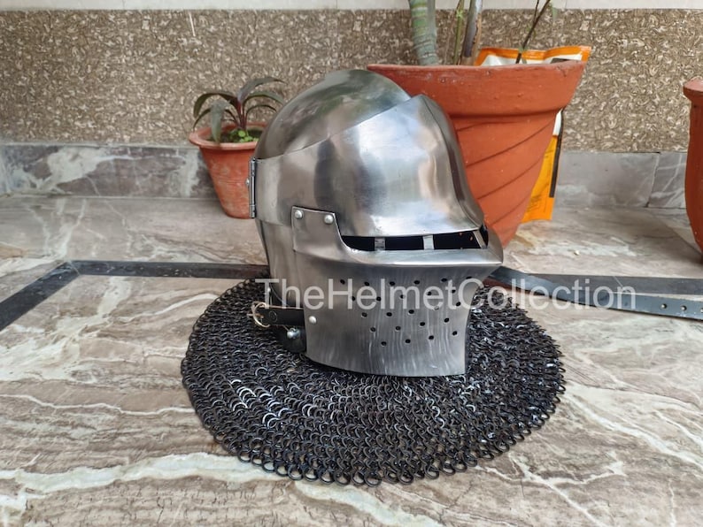 May include: A polished steel helmet with a visor and ventilation holes, resting on a chainmail coif. The helmet has a rounded top and a rectangular visor. The text "TheHelmetCollection" is visible on the image.