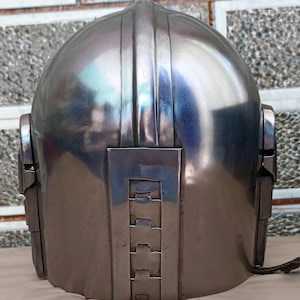 May include: A metallic silver helmet with a rounded top and vertical ridges. The helmet has rectangular side pieces and a series of layered rectangular shapes on the lower back. A dark brown strap extends from the right side.