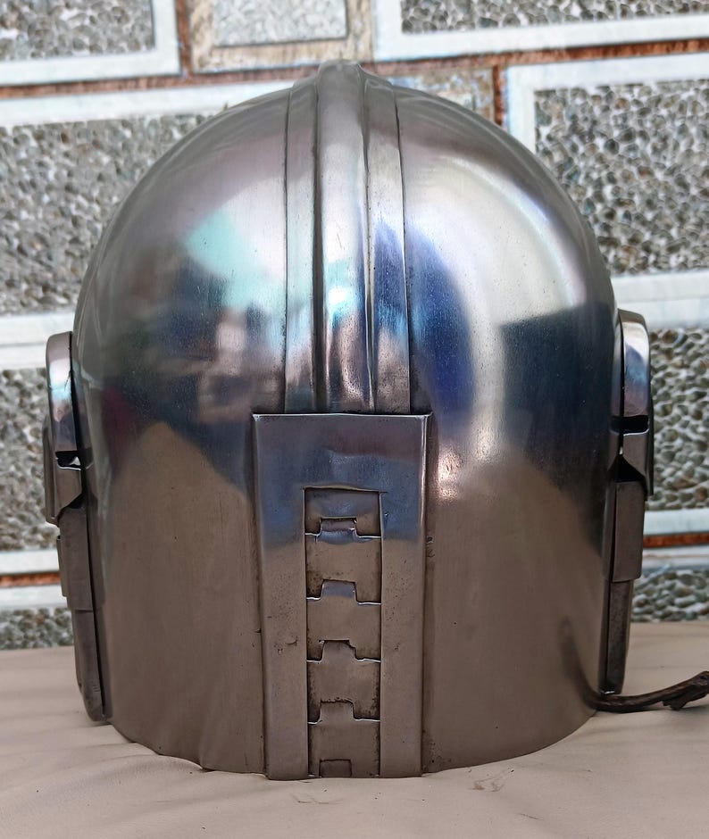 May include: A metallic silver helmet with a rounded top and vertical ridges. The helmet has rectangular side pieces and a series of layered rectangular shapes on the lower back. A dark brown strap extends from the right side.