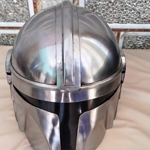 May include: A metallic silver helmet with a black visor. The helmet has a rounded top with vertical ridges and a curved front. The design is reminiscent of a space-themed character.
