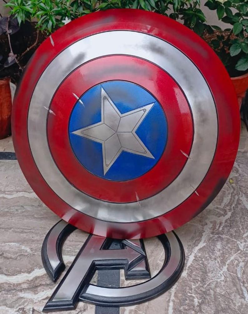 May include: A Captain America shield, featuring a red, white, and blue color scheme. The shield has a circular design with a silver star in the center. The shield is resting on a silver Avengers logo.