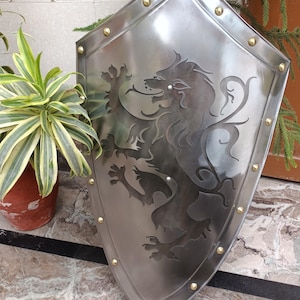 May include: A silver shield featuring a lion emblem. The shield has a rounded top and is edged with brass rivets. The lion is etched into the metal, creating a detailed design. The shield is displayed upright.