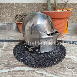 May include: A polished steel helmet with a visor and a chainmail neck guard. The helmet has a rounded top and a series of small holes near the visor. The text "The Helmet Collection" is visible on the image.