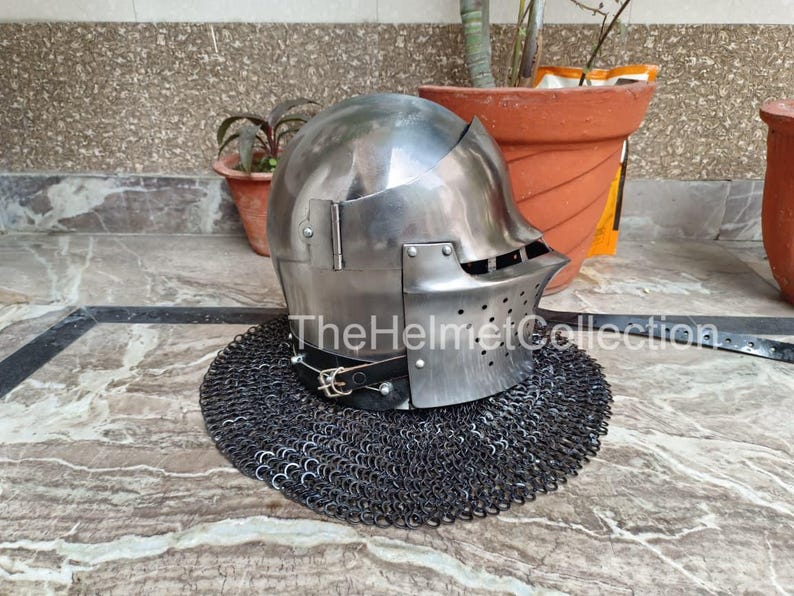 May include: A polished steel helmet with a visor and a chainmail neck guard. The helmet has a rounded top and a series of small holes near the visor. The text "The Helmet Collection" is visible on the image.