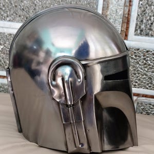 May include: A metallic silver helmet with a visor and distinctive ear-like protrusions. The helmet has a smooth, reflective surface, suggesting a polished metal finish. The design is reminiscent of a science fiction or fantasy theme.