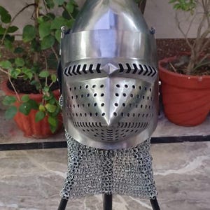 Peut inclure: Casque en m&eacute;tal poli avec une pointe et une protection faciale perfor&eacute;e. Il est dot&eacute; d'une protection de cou en cotte de mailles et est pr&eacute;sent&eacute; sur un support noir. En arri&egrave;re-plan, des plantes vertes en pot.