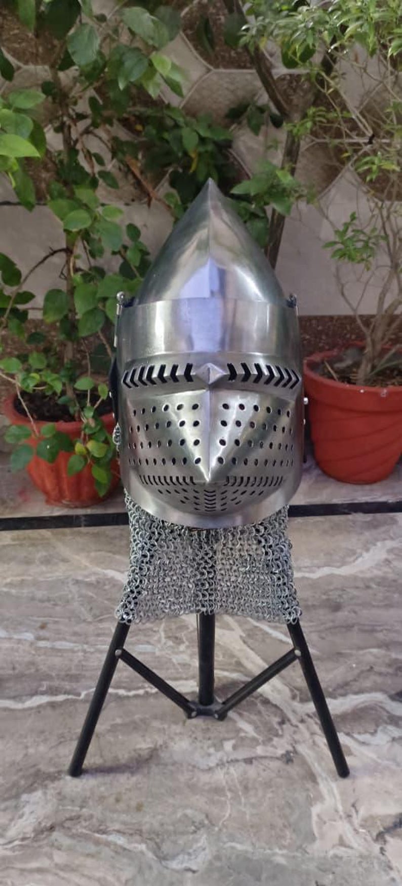 Peut inclure: Casque en m&eacute;tal poli avec une pointe et une protection faciale perfor&eacute;e. Il est dot&eacute; d'une protection de cou en cotte de mailles et est pr&eacute;sent&eacute; sur un support noir. En arri&egrave;re-plan, des plantes vertes en pot.